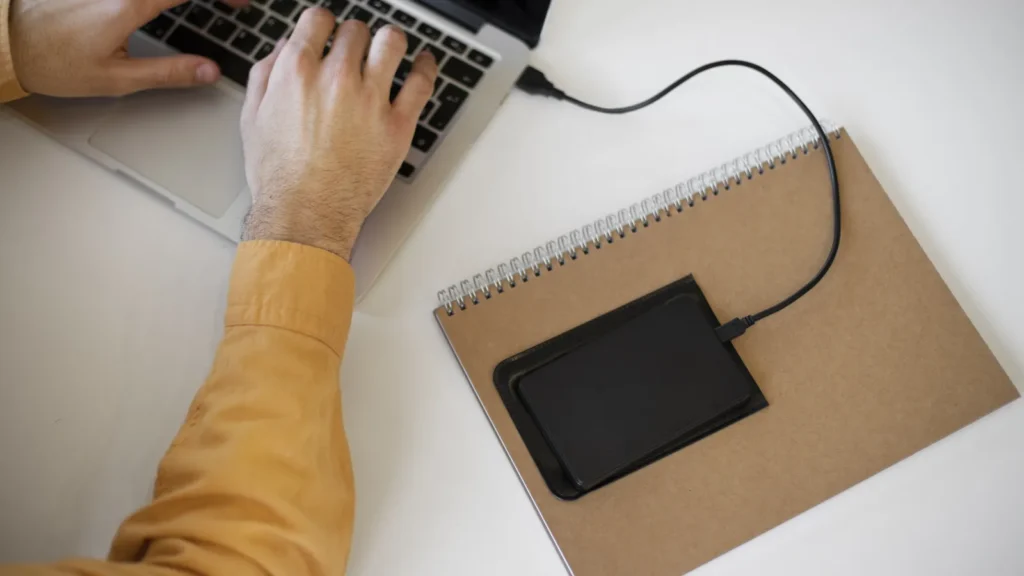 Mani al portatile collegato a un disco esterno appoggiato su un quaderno